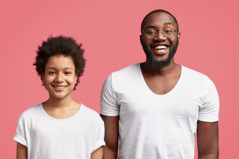 Horizontal shot of happy young African American father stands closely to his little mixed race son, being in good mood, have broad smiles, rejoice seeing guests, isolated over pink background.