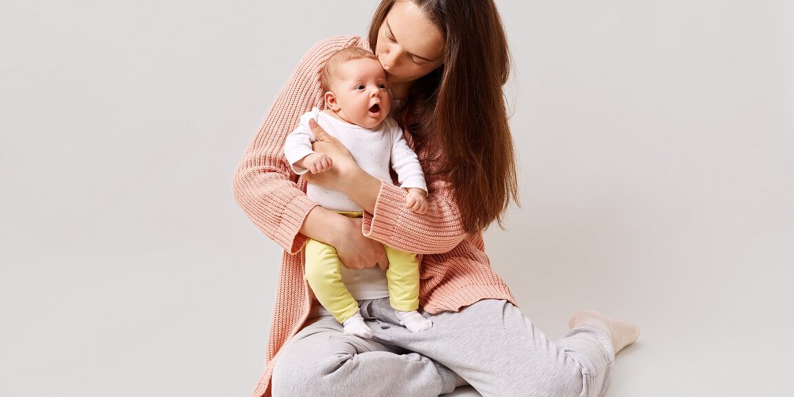 Young beautiful attractive mother holding and kissing newborn baby while sitting on floor, dark haired female wearing casual attire, isolated over white background.