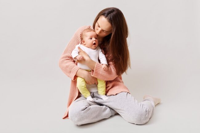 Young beautiful attractive mother holding and kissing newborn baby while sitting on floor, dark haired female wearing casual attire, isolated over white background.